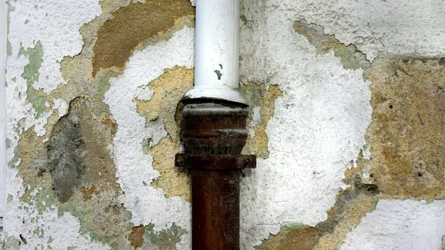 Corroded drain pipe in a plaster wall with water damage around it