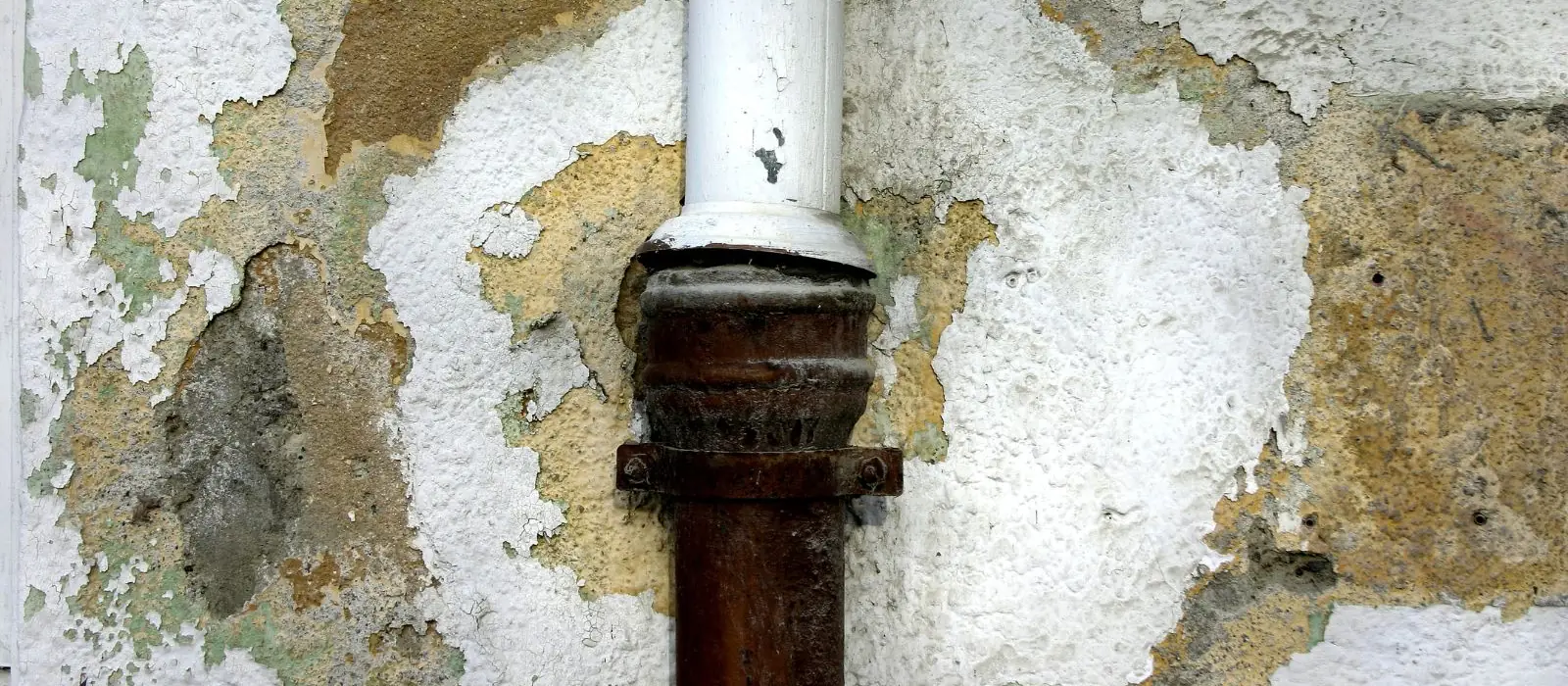Corroded drain pipe in a plaster wall with water damage around it